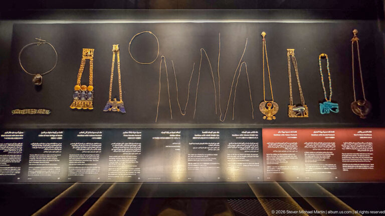 Museum case displaying multiple necklaces, pendants, chains, and colorful amuletic jewelry from Tutankhamun's burial assemblage.