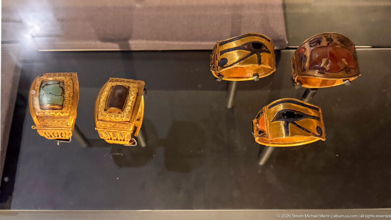 Small gold amulets and jewelry fittings with pale, dark, and reddish inlays arranged in a museum case.