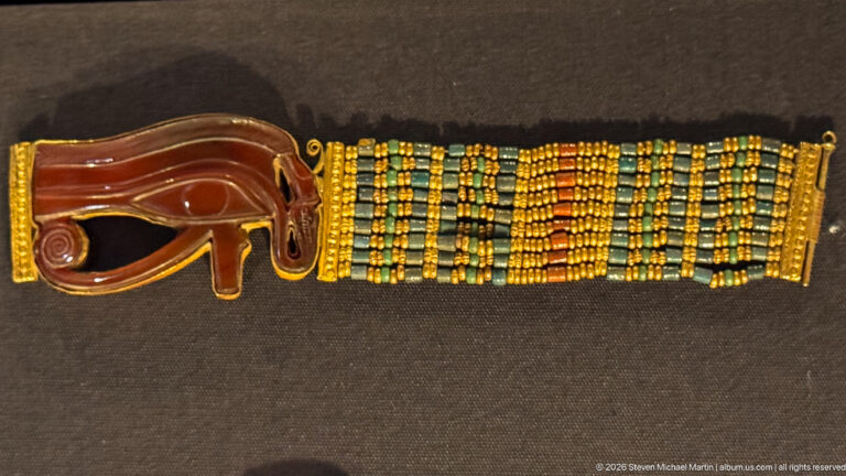 Beaded bracelet with a large reddish Eye of Horus centerpiece and alternating turquoise-blue and gold rows.