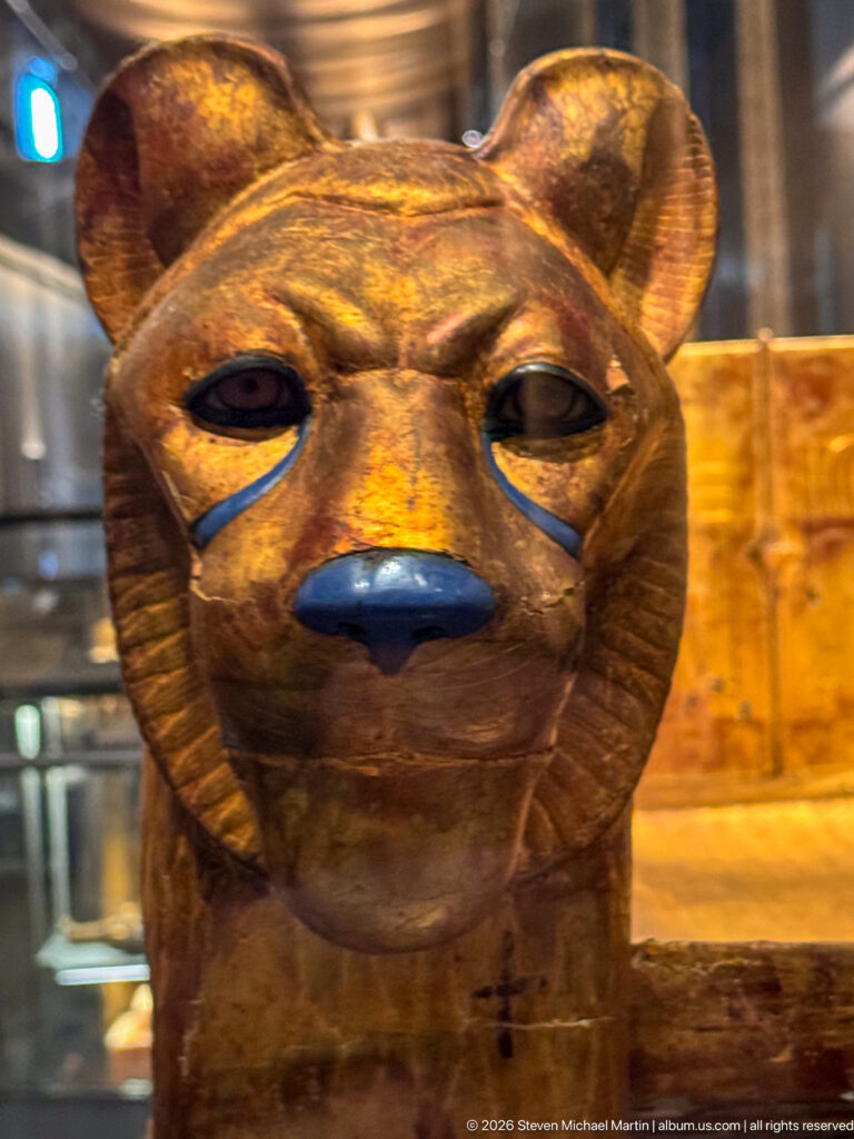 Close frontal view of a gilded feline head with large ears, almond eyes, and blue-painted nose and eye lines.