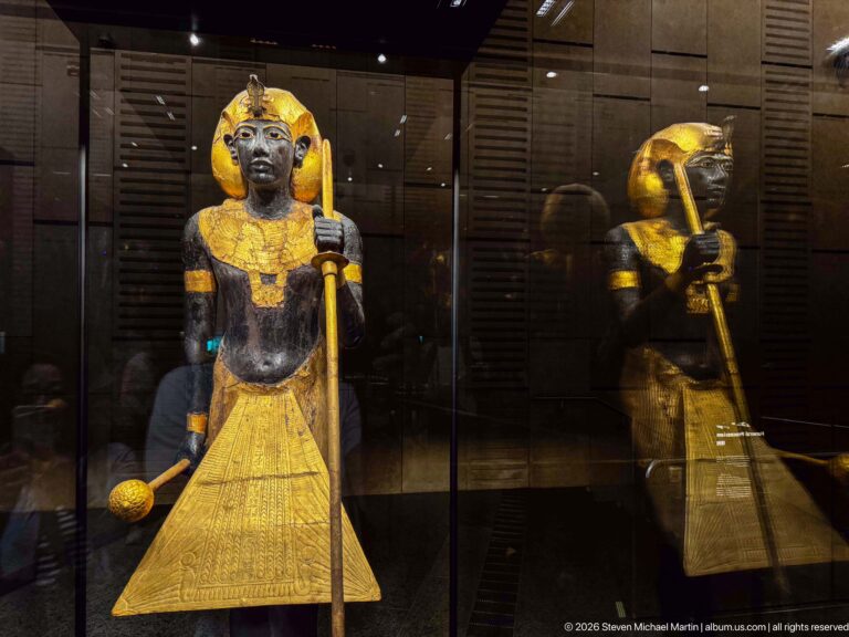 Black-and-gold guardian statue of Tutankhamun with staff and mace beside its reflection in the gallery glass.
