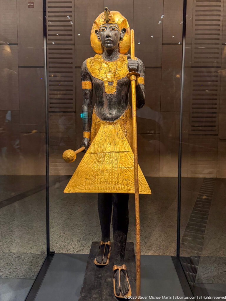 Life-sized black-and-gold guardian statue of Tutankhamun wearing a nemes headcloth and holding a long staff and mace.