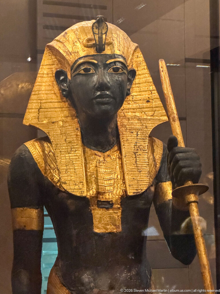 Frontal black-and-gold guardian statue of Tutankhamun in nemes headcloth holding a long staff and mace.