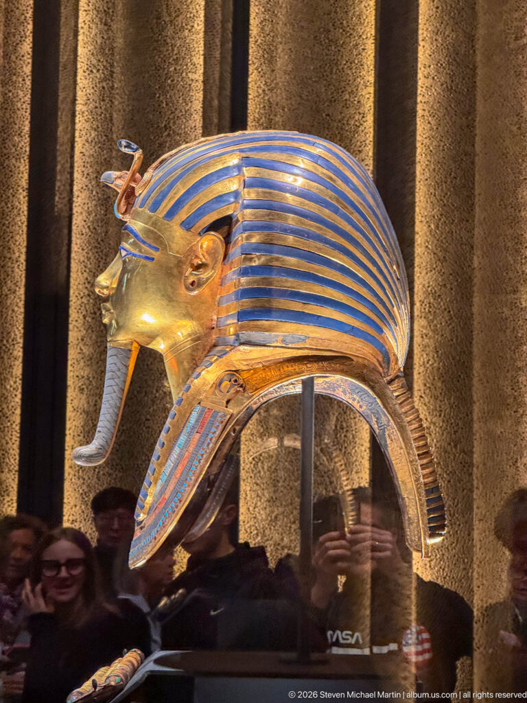 Left profile of Tutankhamun’s solid-gold burial mask with striped nemes, brow emblems, false beard, and inlaid collar.