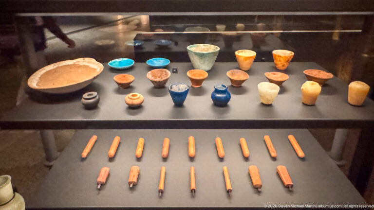 Small bowls, cups, jars, and orange rod-like implements arranged in two rows inside a display case.