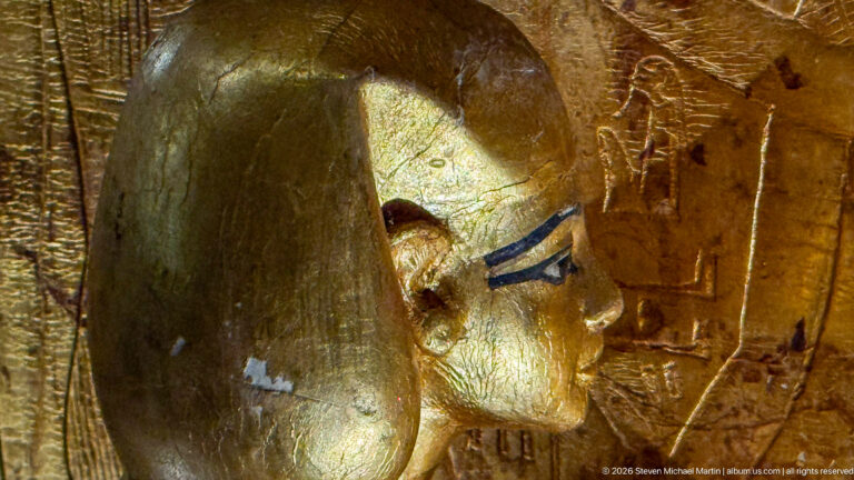 Profile of a gilded Egyptian face with blue eye cosmetics beside incised hieroglyphic decoration.