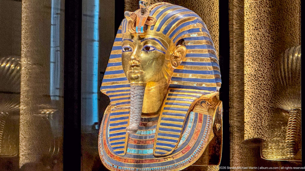 Three-quarter view of Tutankhamun’s solid-gold burial mask with blue-striped nemes, false beard, brow emblems, and inlaid collar.