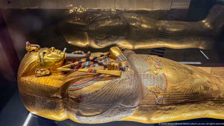 Oblique close view of a gilded anthropoid coffin with royal face, crossed regalia, and dense colored patterning.