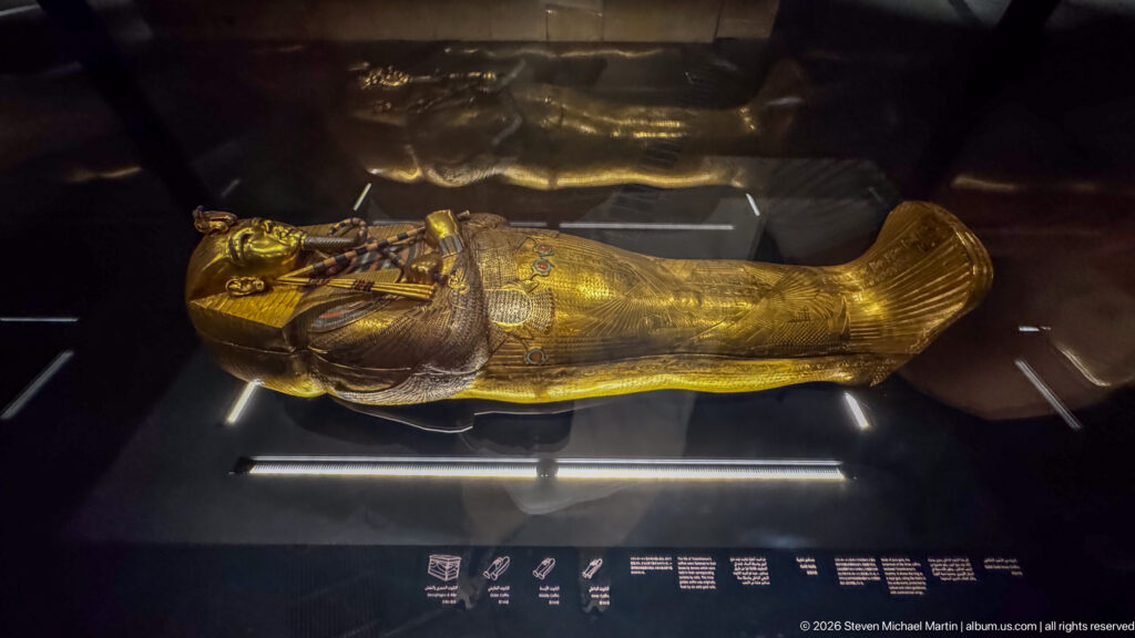 Full-length gilded anthropoid coffin lying horizontally in a dark museum case.