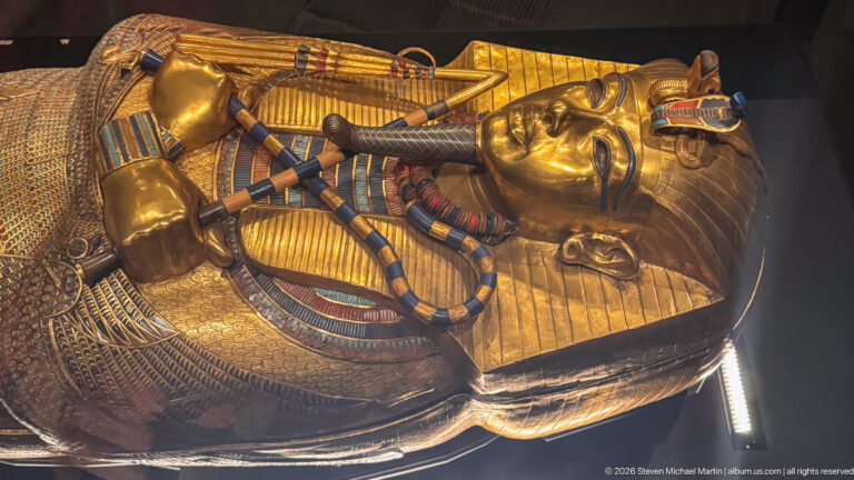 Oblique close view of gilded royal coffin with face, broad collar, crossed regalia, and colored inlay.