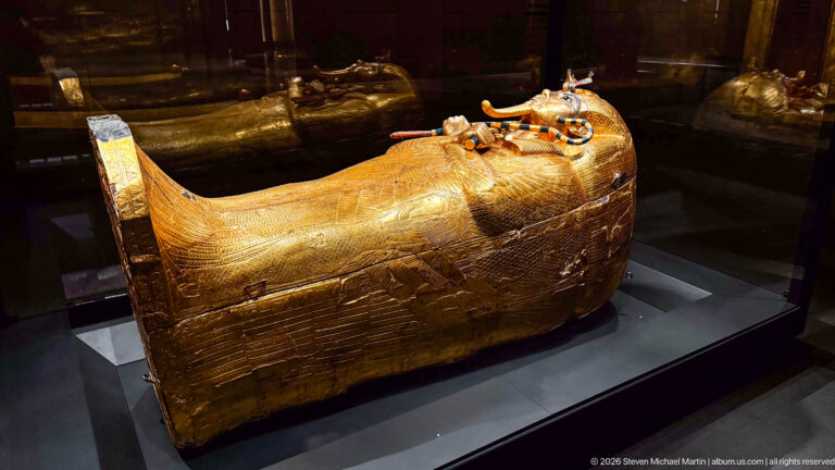 Full-length gilded anthropoid coffin of Tutankhamun lying in a dark glass display case.