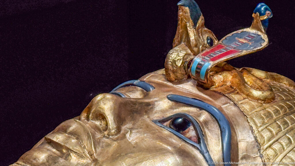 Close gilded royal face with cobra and vulture emblems, blue inlay around the eyes, and headdress details.