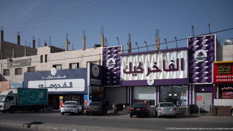 Purple-and-white Arabic furniture storefront with parked cars and an unfinished building structure rising behind it.