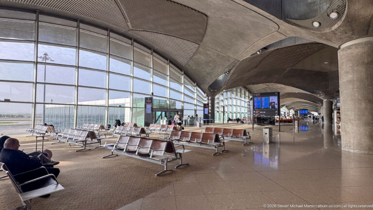 Airport waiting area with rows of seats beneath sculptural concrete roof modules and tall glass walls.