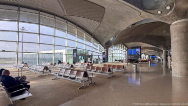 Airport waiting area with rows of seats beneath sculptural concrete roof modules and tall glass walls.