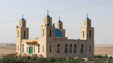 Compact pale-stone mosque with blue dome, four square corner towers, green doors, and loudspeakers in a dry landscape.