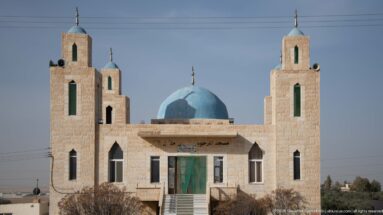 Small pale-stone mosque with bright blue dome, four corner towers, green openings, and mounted loudspeakers.