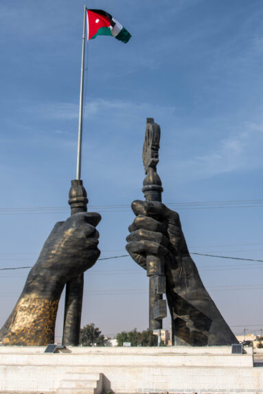 Large sculptural hands flank a central pedestal beneath a tall Jordanian flag flying against the blue sky.
