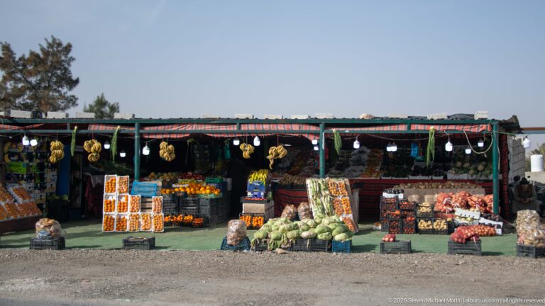 Open roadside produce stand with stacked watermelons, melons, oranges, and other fruits beneath a shaded canopy.