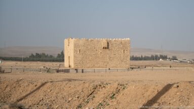 Distant side view of a compact crenellated stone fort standing alone on open dry ground.