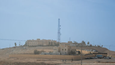 Pale hilltop building complex with a tall communications mast above graded dry terrain beside a road.