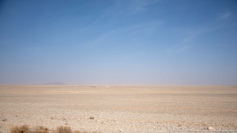 Wide pale stony desert plateau stretching to a hazy horizon beneath a clear blue sky.