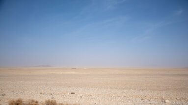 Wide pale stony desert plateau stretching to a hazy horizon beneath a clear blue sky.
