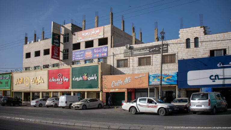 Arabic-signed storefronts, parked vehicles, and unfinished upper floors along a commercial road in Amman.