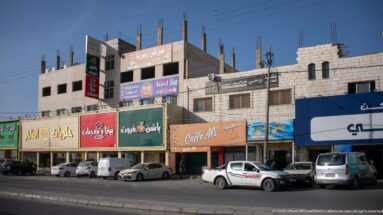 Arabic-signed storefronts, parked vehicles, and unfinished upper floors along a commercial road in Amman.