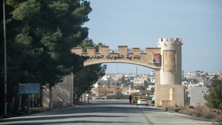 Stone-colored roadside entrance arch with Arabic inscription and crenellated detailing spanning a road toward dense housing.