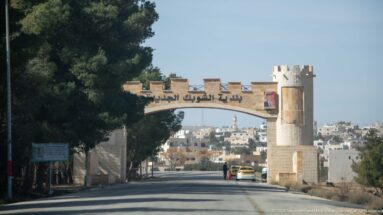 Stone-colored roadside entrance arch with Arabic inscription and crenellated detailing spanning a road toward dense housing.