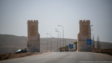 Road passes between two tall crenellated stone-clad gateway towers with streetlights and dry hills beyond.