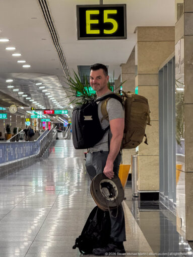 Traveler with large backpack and hand luggage stands directly beneath an illuminated E5 sign in an airport terminal.