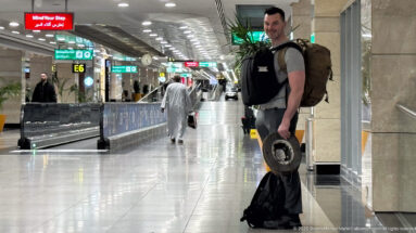Traveler with a large backpack and black bag stands in a bright airport concourse near gate E6.