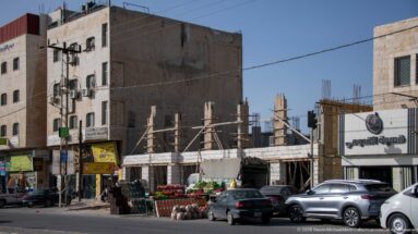 Roadside produce stalls, parked cars, shopfronts, and partly unfinished concrete buildings along an Amman street.