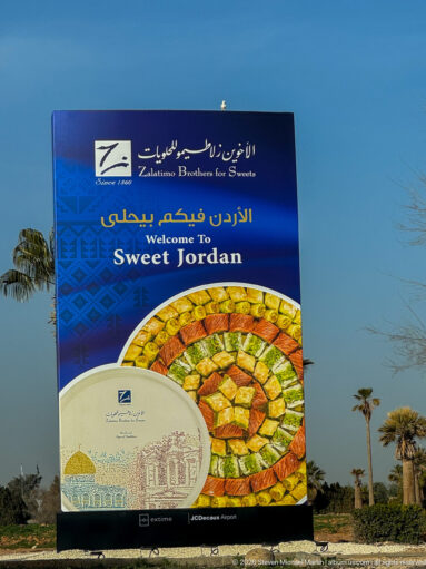 Blue bilingual welcome billboard advertising Zalatimo Brothers for Sweets with a large circular display of pastries.