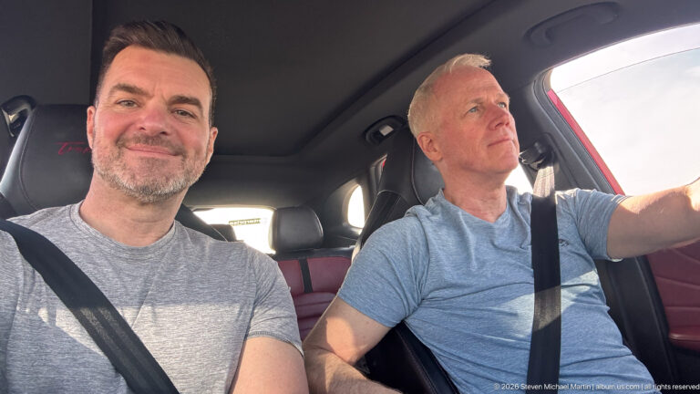 Passenger smiles at the camera while the driver looks ahead inside a sunlit car during a Jordan road trip.