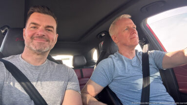 Passenger smiles at the camera while the driver looks ahead inside a sunlit car during a Jordan road trip.