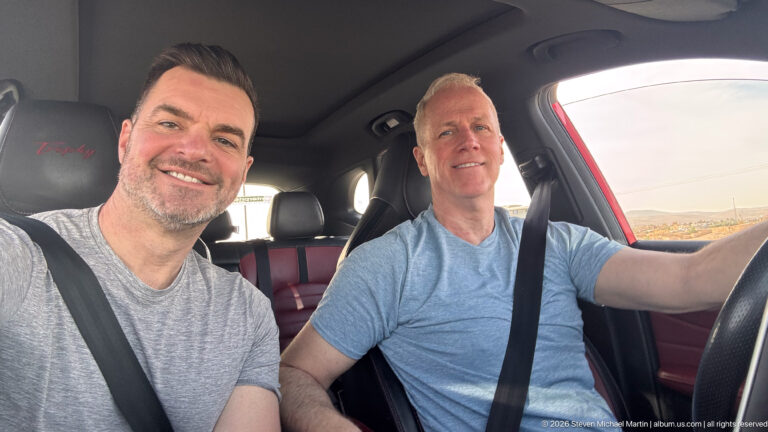 Two smiling men wearing seat belts sit in the front of a car, one driving and one taking the selfie.