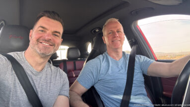 Two smiling men wearing seat belts sit in the front of a car, one driving and one taking the selfie.
