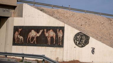 Concrete highway wall with a mosaic-style panel of camels, circular emblem, and smaller painted symbols.