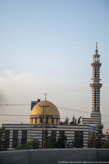 Modern mosque with a broad gold dome and tall horizontally banded minaret seen across roadside barriers in Amman.