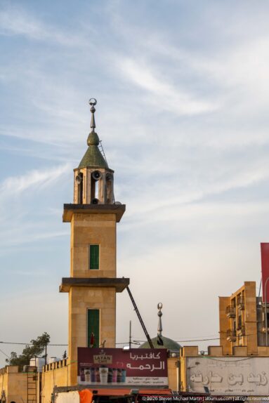 Square mosque tower with open upper pavilion and finial rising above roadside shops and signs in Amman.