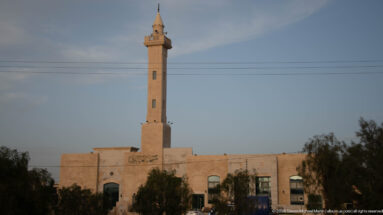 Pale-stone neighborhood mosque with a tall slender minaret, Arabic façade inscription, and trees at dusk.