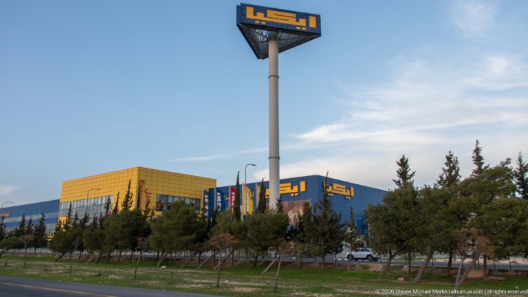 Blue-and-yellow IKEA store and tall pole sign behind roadside trees and landscaped grounds in Amman.