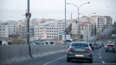 Cars travel on a multi-lane Amman roadway bordered by concrete barriers, dense hillside housing, streetlights, and a distant minaret.