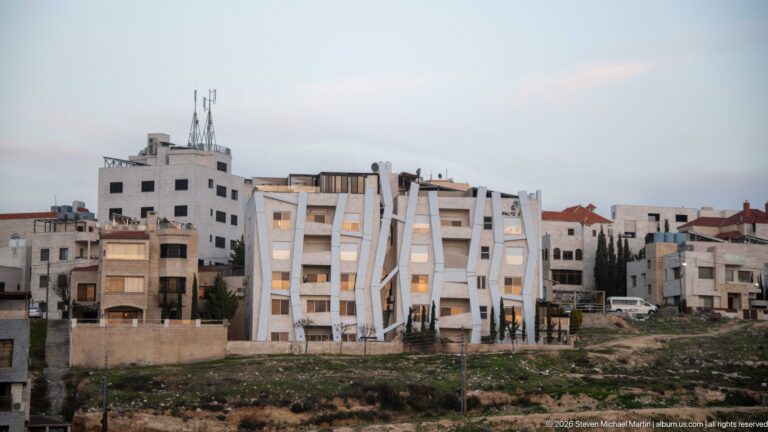 White Amman apartment building with angular projecting frames and diagonal façade elements among pale residential blocks.