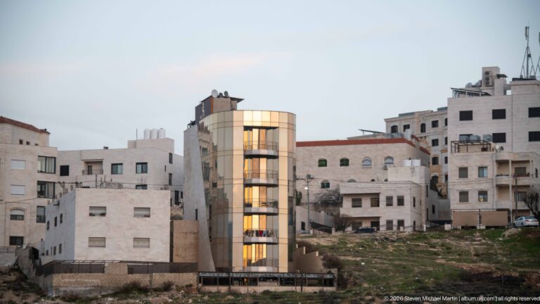 Amman apartment building with a glowing curved glass vertical bay amid pale stone residences and undeveloped ground.