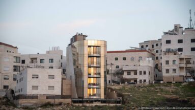 Amman apartment building with a glowing curved glass vertical bay amid pale stone residences and undeveloped ground.