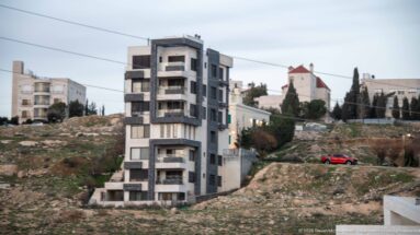 Narrow modern apartment building with stacked balconies on a rocky Amman slope, surrounded by older housing and open ground.